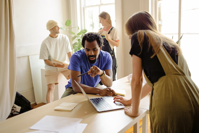 A man talking to a young woman in front of a laptop, with 2 house painters in the background.